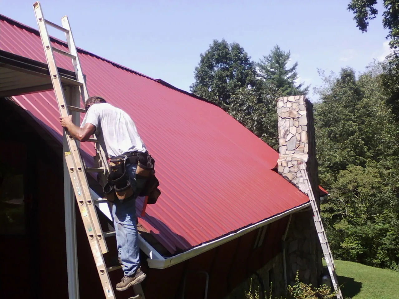 Roofer on a ladder beside a red metal roof and stone chimney — Soffit & Fascia Repair by Roofing Brothers in Harrodsburg